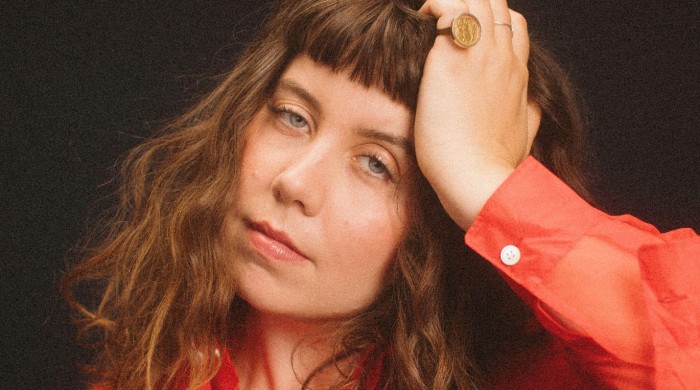 A woman wearing a red shirt with her hand rested on her head. She has a large gold coin ring on her pinky finger. She has a curly brown bob with a fringe